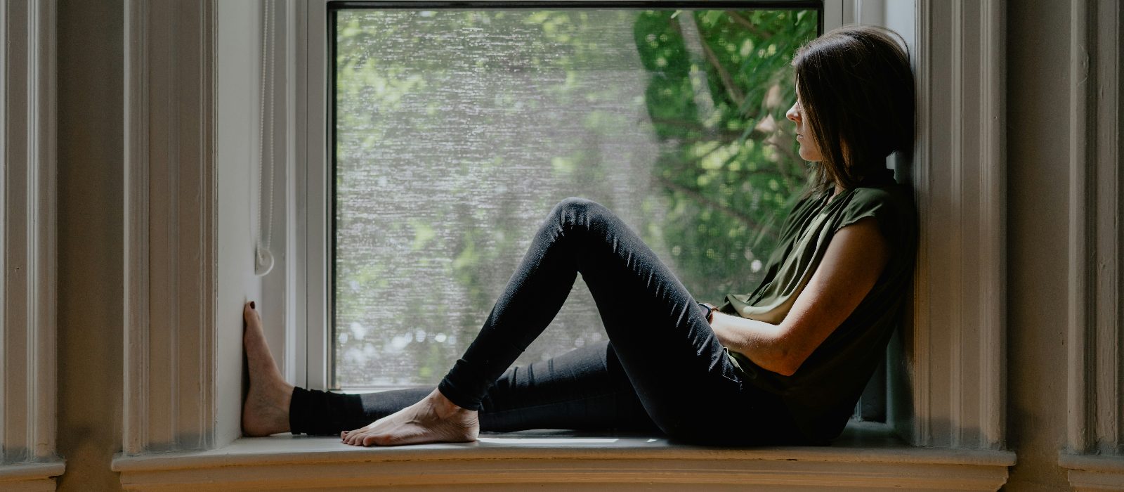 A woman sitting in a windowsill, looking out at green trees