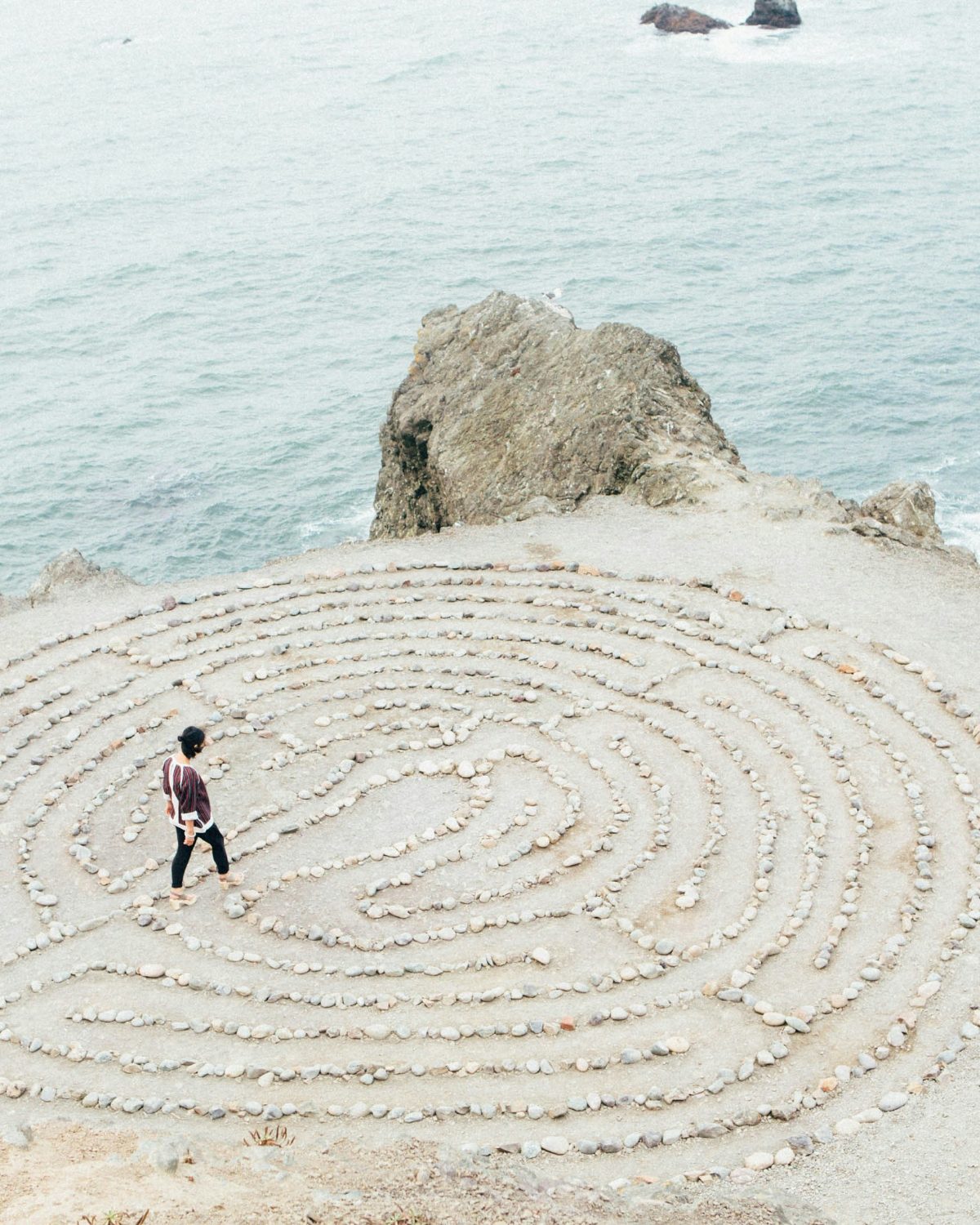 A person walking a stone spiral labyrinth on a coastal cliff above the ocean