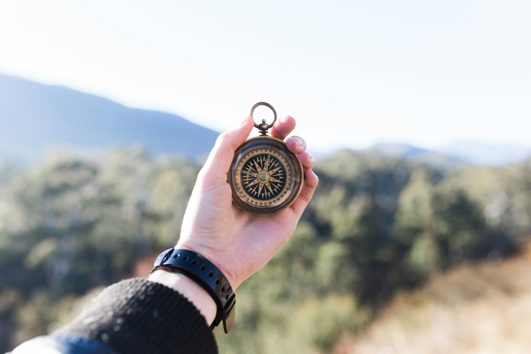 A hand holding a brass compass against a mountain and forest backdrop
