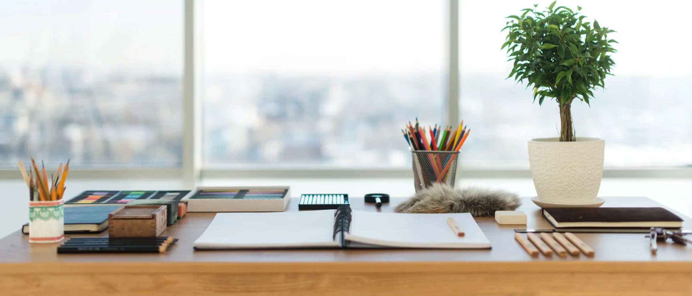 A wooden desk with an open notebook, pencils, keys, and a small potted plant in front of a floor-to-ceiling window